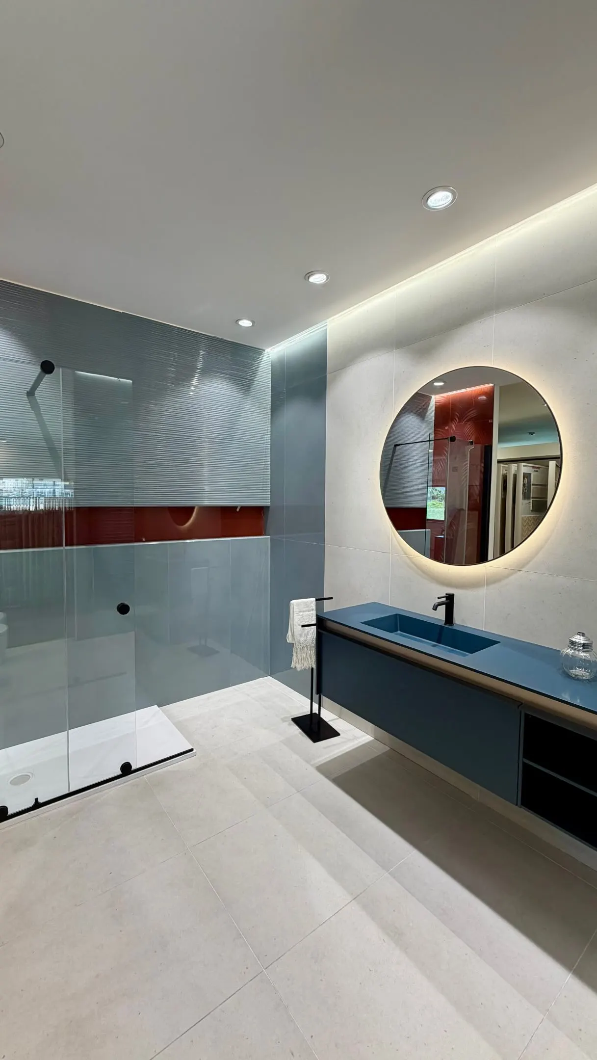 Bagno con mobile sospeso e lavabo integrato blu petrolio, miscelatore nero, specchio tondo retroilluminato e doccia walk-in con parete in vetro