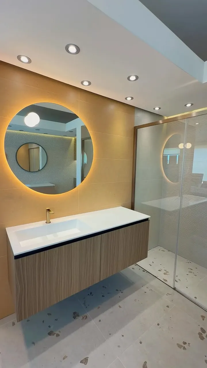 Bagno con specchio tondo retroilluminato su parete beige, mobile sospeso effetto legno con lavabo integrato bianco e miscelatore oro, box doccia a fianco
